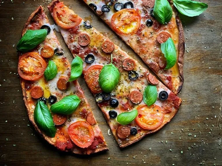 Pizza with pepperoni, tomato slices, black olives, and fresh basil leaves on a wooden surface, sliced into four pieces.